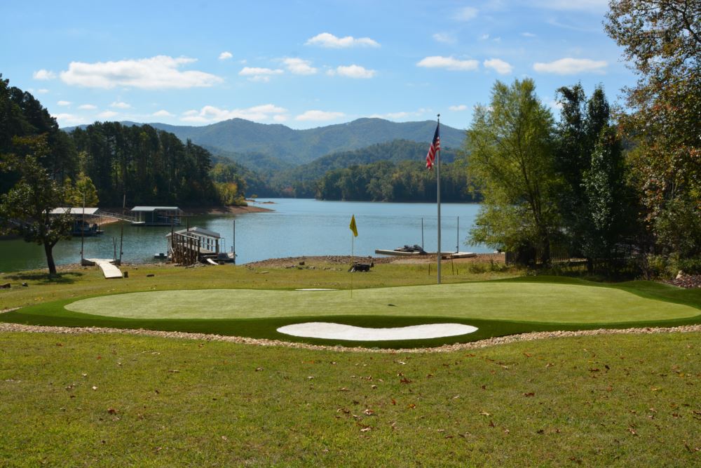 an artificial grass putting green with an artificial grass sand bunker and American Flag, lake, and mountains in the background