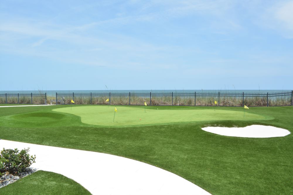 Synthetic turf putting green with multiple flagsticks and a winding walkway with a turf bunker with a fence and blue sky in the background.