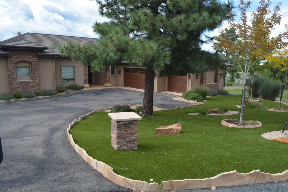 A front yard with an artificial grass lawn installed surrounded by a rock border and with trees growing around it