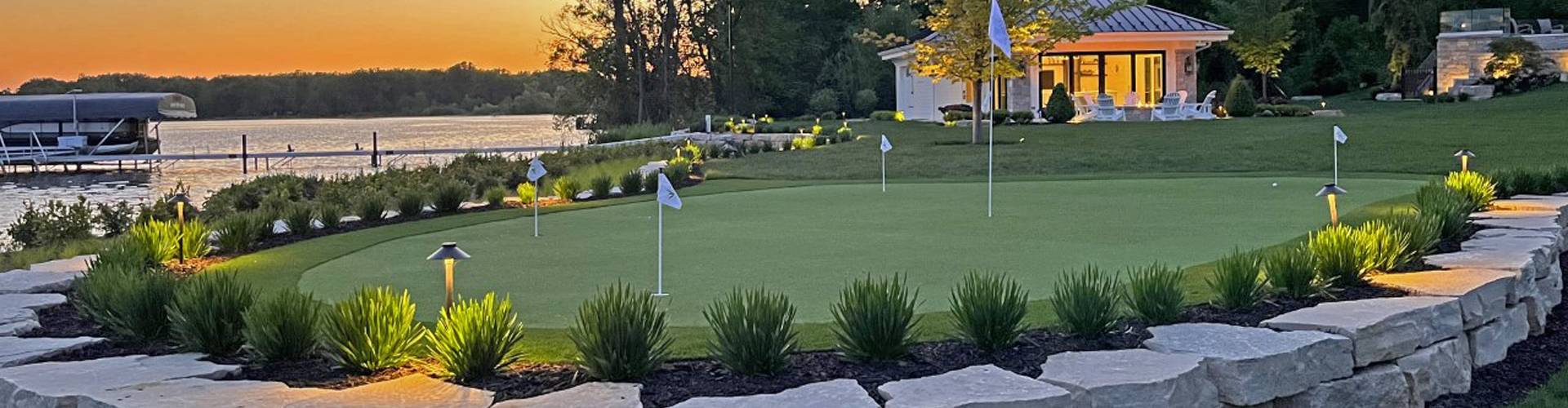 Southwest Greens putting green at sunset on a private lakehouse estate with five flag holes, landscape lighting, stone retaining wall, and boat dock.